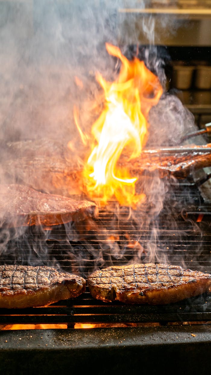 Sizzling steaks cooking on an open flame barbecue with smoke and fire.